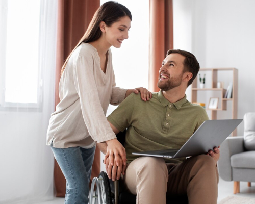 Smiling caregiver assisting a man in a wheelchair using a laptop at home, representing compassionate in-home support by an NDIS Provider.