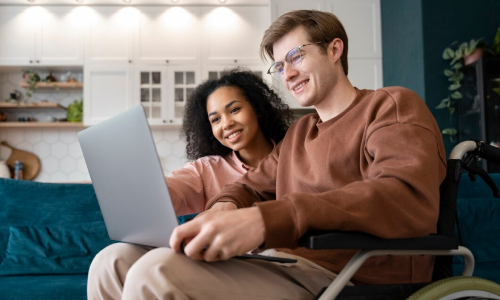 Happy couple using a laptop together at home, with one person in a wheelchair, representing inclusive living and supportive care from an NDIS Provider.