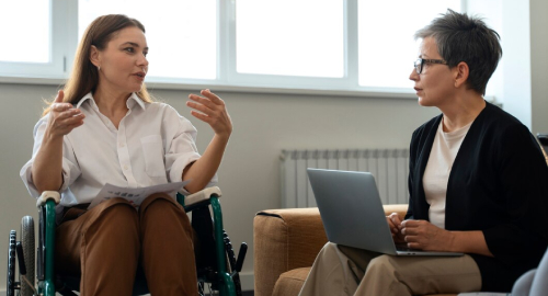 A woman in a wheelchair actively speaking and gesturing with her hands while holding papers, engaged in a discussion with a professional woman holding a laptop. They are sitting face-to-face in a bright, modern room. NDIS Support Service Provider.