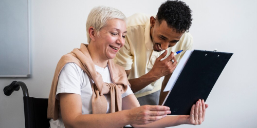 An elderly woman in a wheelchair smiling happily while holding a clipboard together with a young male caregiver who is pointing at the document with a pen. They appear engaged and cheerful.