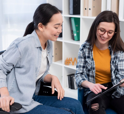 Two women sitting together in a modern office, smiling and looking at a tablet; one is pointing at the screen while the other leans in, engaged in the discussion. Shelves with binders and decor are visible in the background.