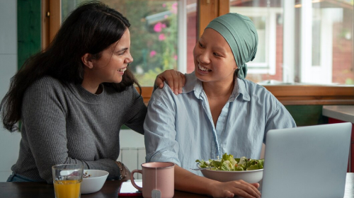 A woman gently rests her hand on another woman’s shoulder as they sit at a table with food and a laptop; the second woman, wearing a headscarf, smiles back. A glass of orange juice and a salad bowl are on the table, and daylight streams through a nearby window.