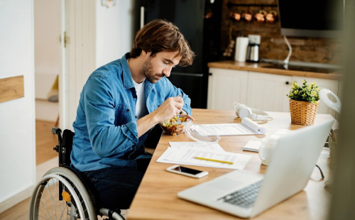 A man using a wheelchair sits at a kitchen table eating a meal from a container while looking down; papers, a smartphone, and an open laptop are spread out in front of him, with a cozy kitchen in the background.