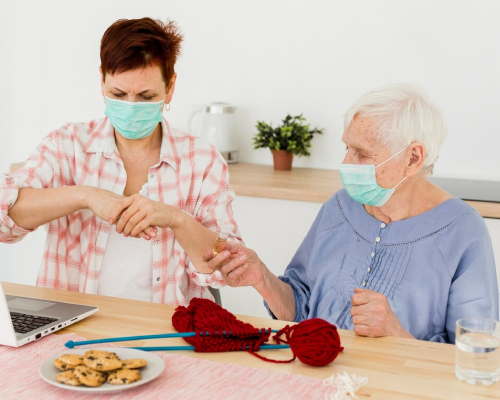 A younger woman wearing a face mask helps an elderly woman apply hand sanitiser at a table with knitting supplies, cookies, and a laptop nearby.