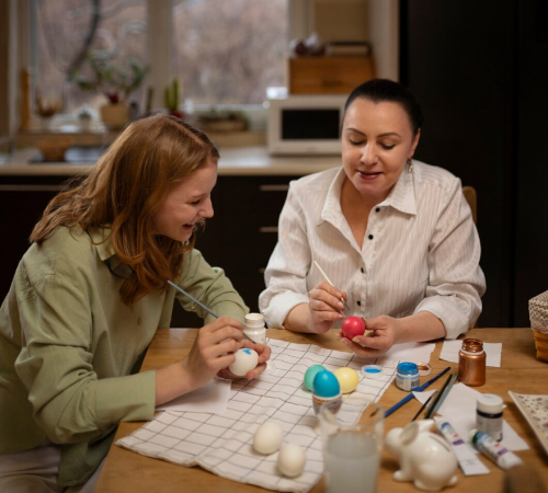 An adult woman and a child sit at a table painting colorful eggs together, smiling and engaged in a craft activity.