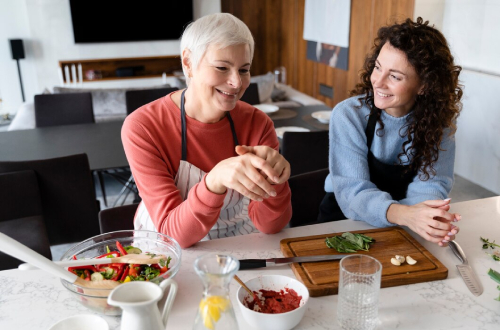 Two women in a modern kitchen smile and chat while preparing food together. One chops herbs on a cutting board while the other mixes ingredients in a bowl, with vegetables and utensils spread across the counter.