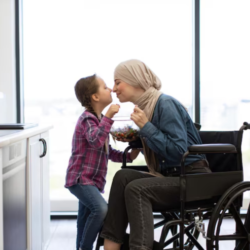In a bright kitchen, a woman wearing a headscarf sits in a wheelchair while a young girl affectionately feeds her from a bowl. The moment feels warm and personal, symbolizing everyday care and family support.