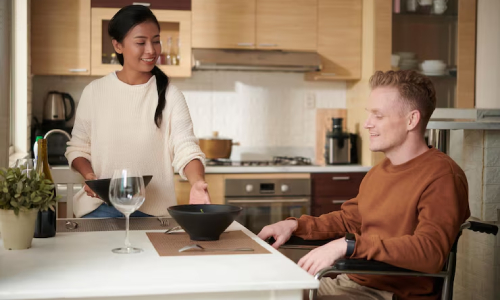 In a modern kitchen, a woman serves a meal to a man seated in a wheelchair. Both are smiling, suggesting a positive and supportive environment where domestic tasks are shared, aligning with the idea of reliable household assistance.